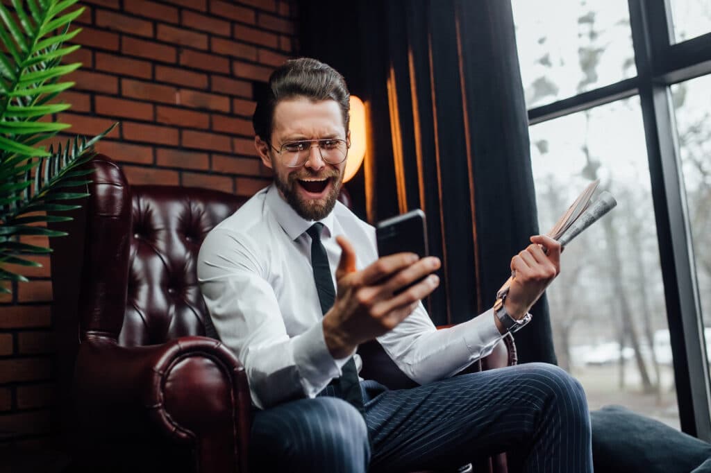 handsome businessman with candid emotions holding his smartphone and newspaper while sitting on armchair Cómo son las casas de apuestas Texas